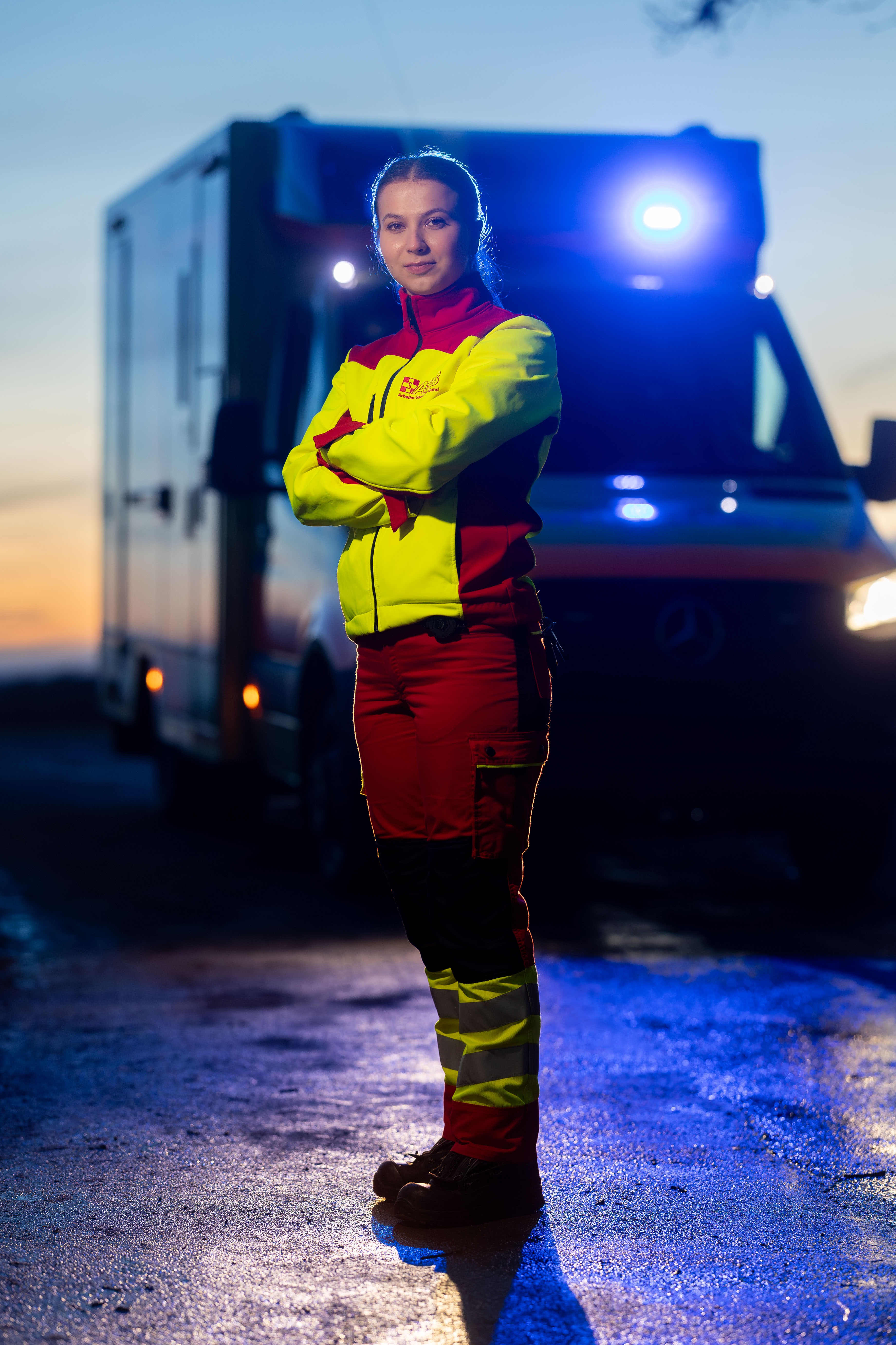 Notfallsanitäterin vom ASB Neckar-Alb vor einem Rettungswagen mit eingeschalteten Blaulicht, die Situation ist in der Abenddämmerung